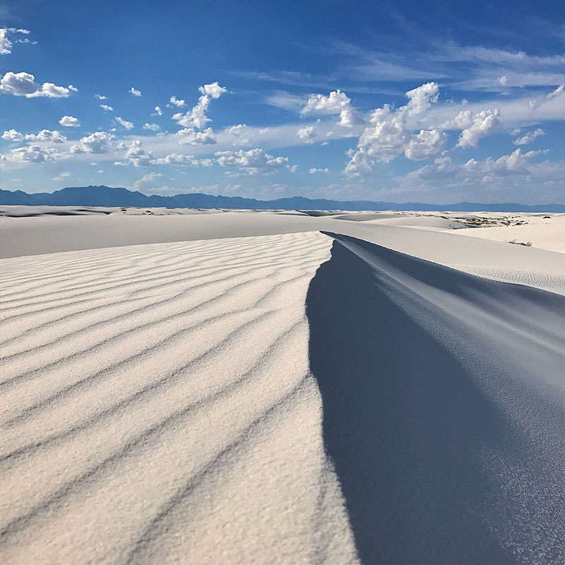 A rippled white sand dune ridge casting a long shadow under a blue sky with scattered clouds
