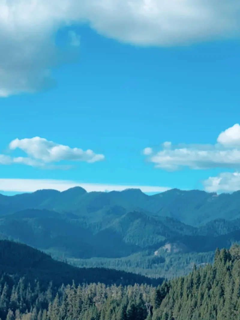 Big white clouds in a blue sky above layered forested mountain ridges