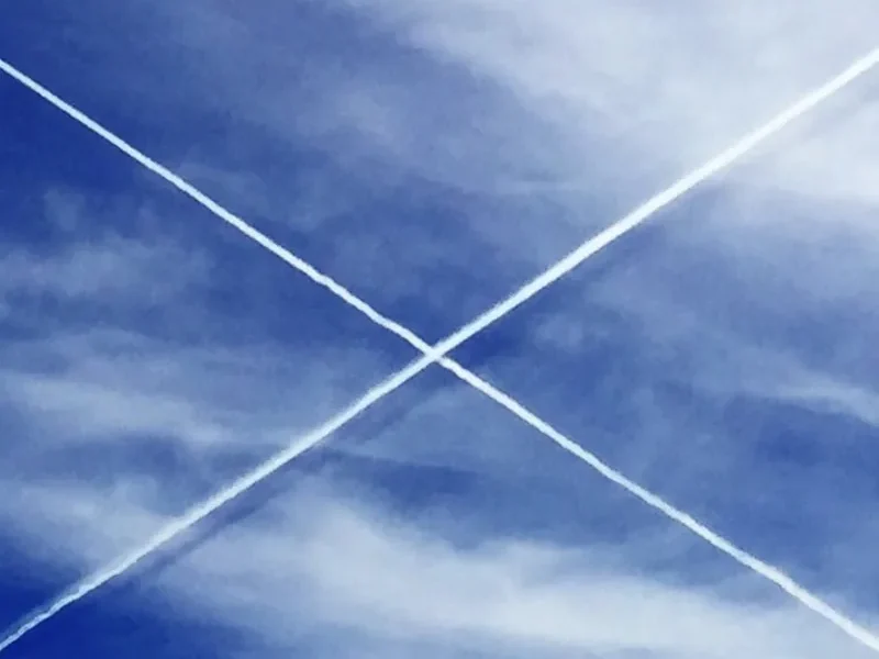 Two white jet contrails crossing in an X across a deep blue sky with wispy clouds
