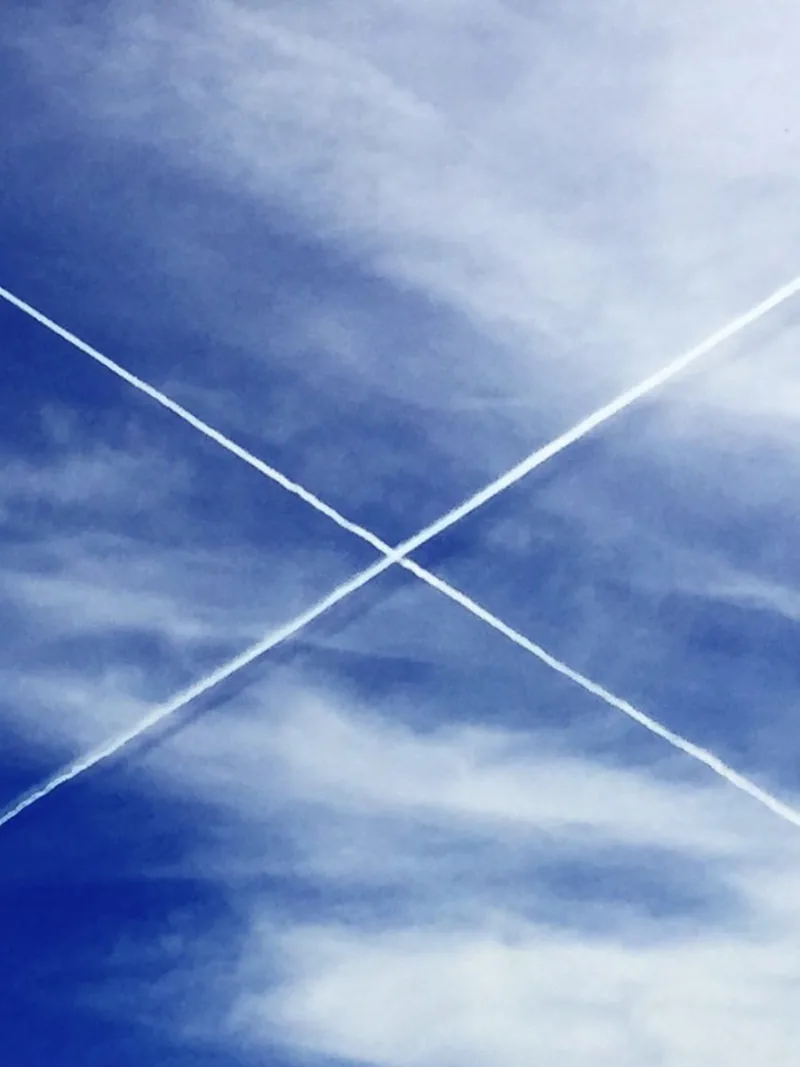 Two white jet contrails crossing in an X across a deep blue sky with wispy clouds