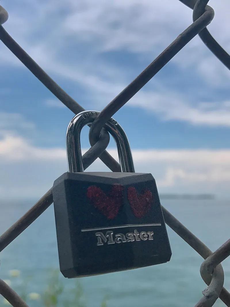 A black padlock locked to a chain-link fence with the ocean and cloudy sky behind it